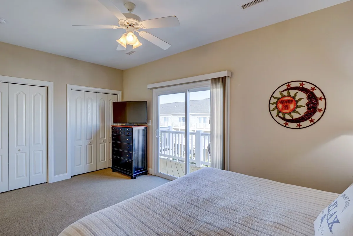 A calm, serene bedroom featuring a bed and dresser, with a sliding glass door that opens to what appears to be an exterior view. The influx of natural light hints at a connection to the outdoors, suggesting access to a private deck or balcony where guests can enjoy coastal sunrises.