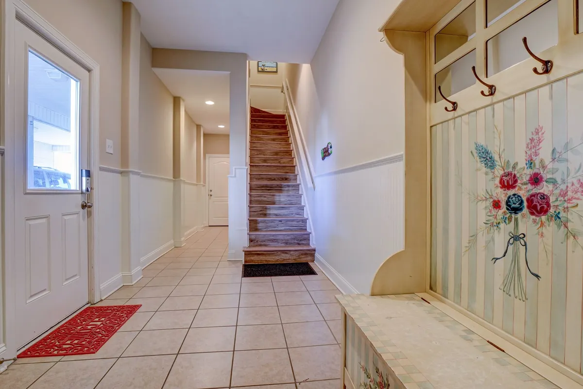 A welcoming entryway inside the home, featuring a staircase, a decorative coat rack, a floral-design bench, and the entry door. The area is functional and inviting, capturing the essence of arriving and settling in.