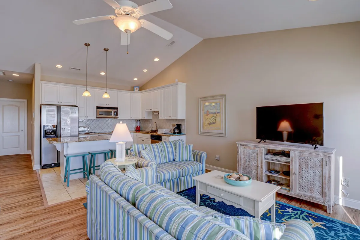 A vibrant view of the open-plan living space, including a striped sofa, kitchen island, TV unit, and dining area. The layout supports group activities such as game nights, movie evenings, and shared meals, with sunlight streaming in from generous windows.