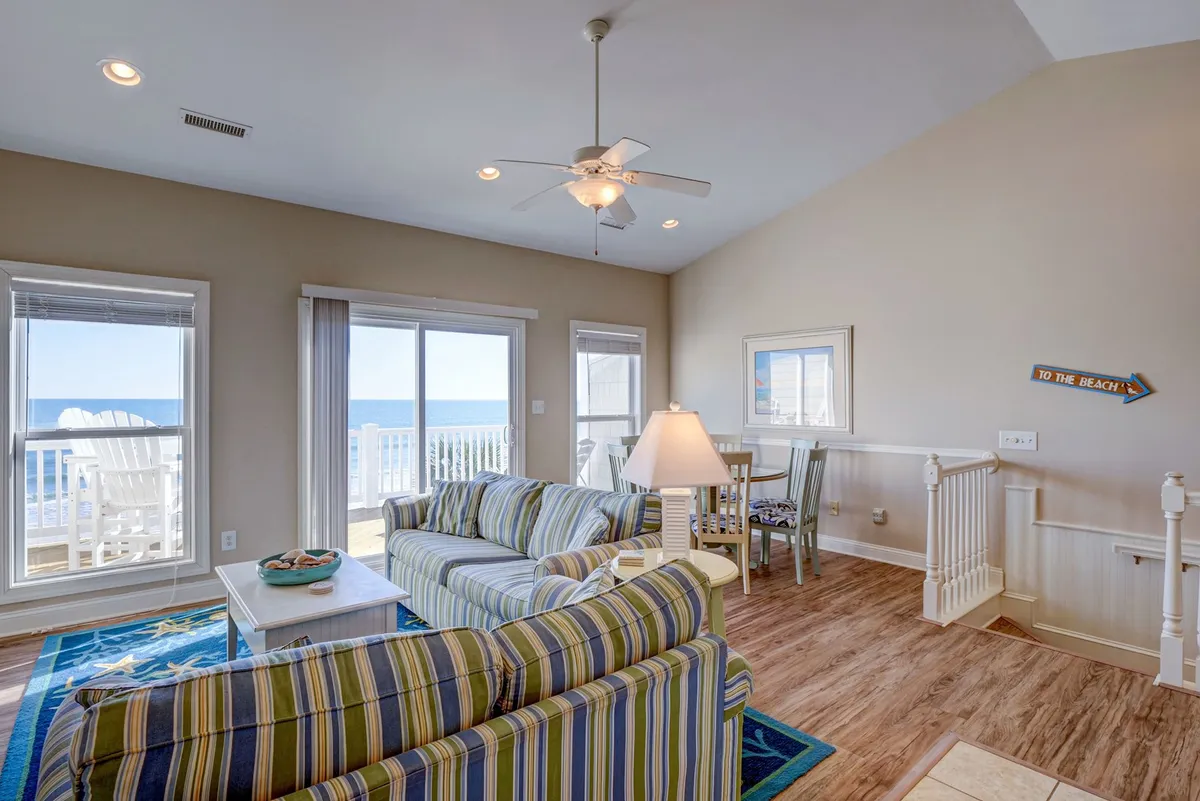 A bright living space featuring a striped sofa positioned to take in expansive ocean views through large windows. Beach-themed artwork and a welcoming sign add to the coastal charm, creating a seamless connection between indoor comfort and the vibrant island atmosphere beyond.