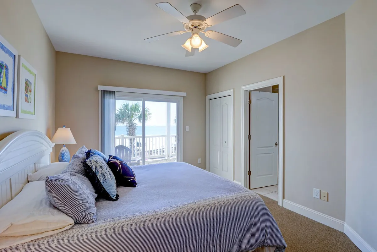 A serene bedroom featuring a bed with decorative pillows and a window overlooking palm trees. The setting is airy and tranquil, with soft natural light filling the space. A ceiling fan and subtle coastal decor create a relaxing atmosphere.