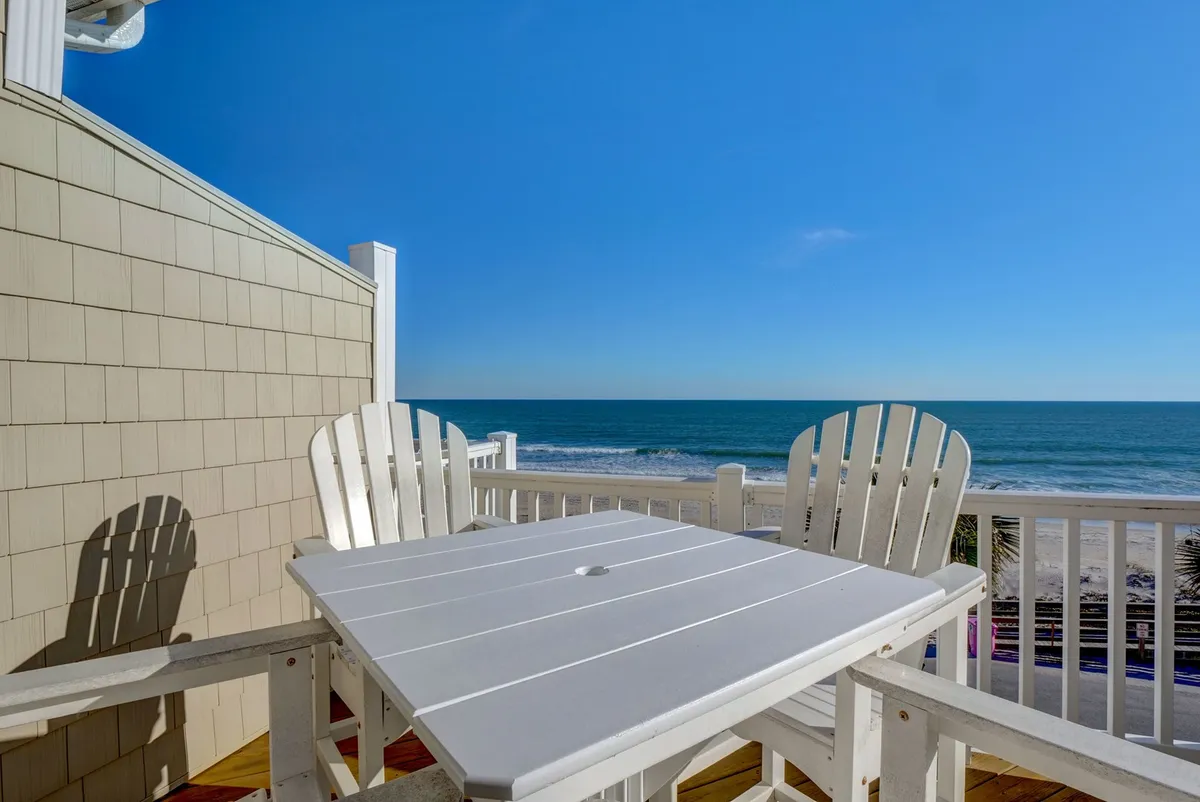 A white patio table with adjoining chairs set on an outdoor terrace, overlooking a breathtaking ocean view. The setting is tranquil yet welcoming, perfect for casual outdoor dining or friendly gatherings.